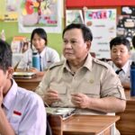 Man in a khaki uniform seated at a desk in a classroom surrounded by students in white shirts and ties